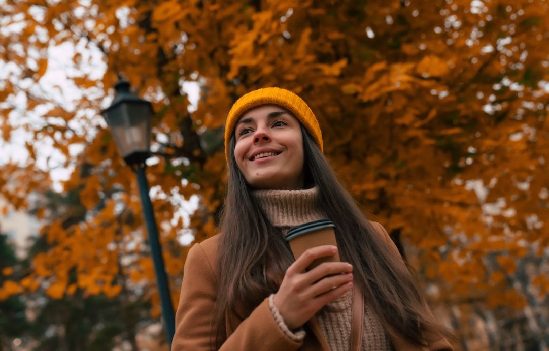 Woman enjoying coffee outdoors in autumn, wearing a yellow beanie, with fall leaves in the background.