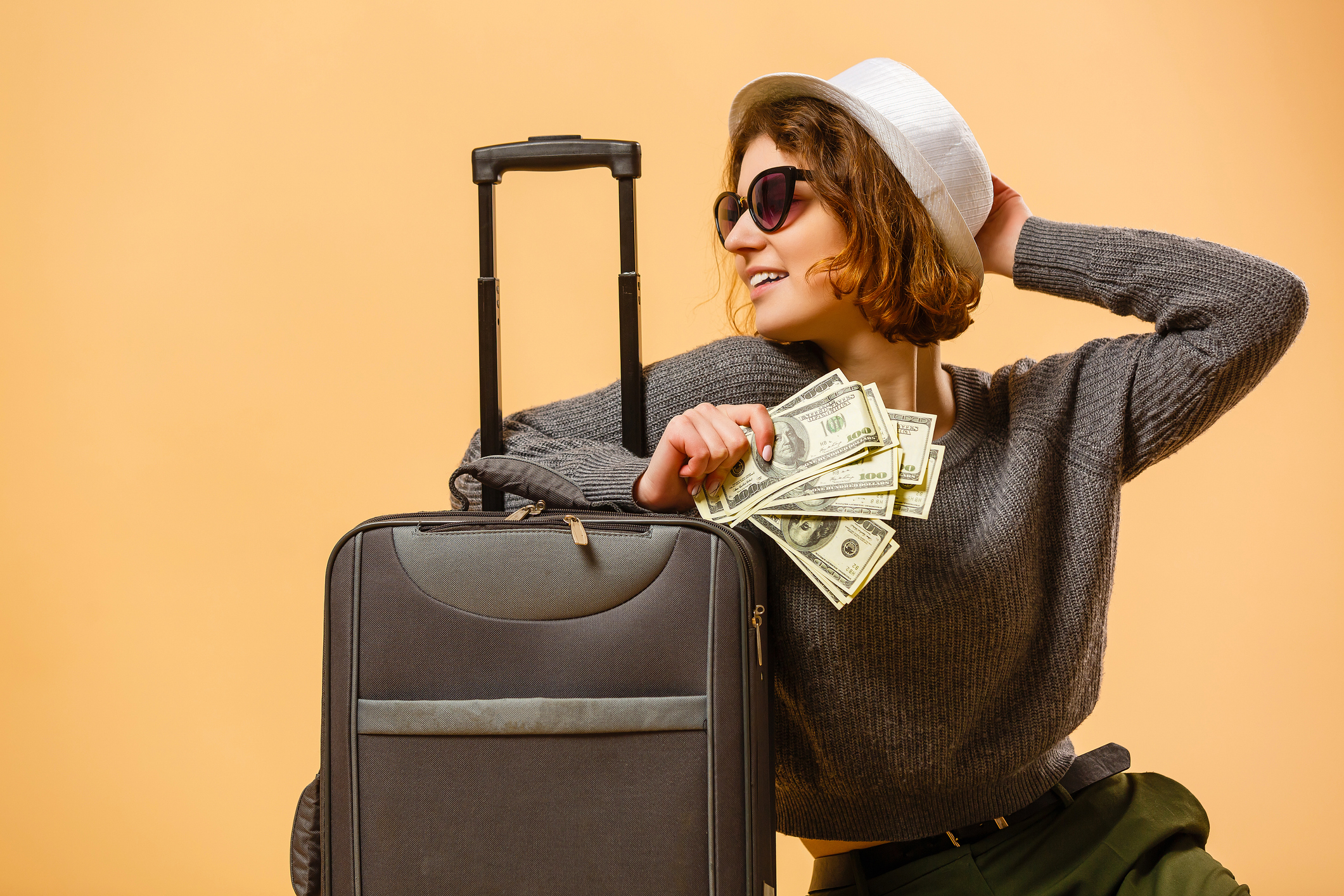 Tourist woman wearing casual clothes, sunglasses, and a hat. Leaning on black suitcase, holding cash in front of yellow background.